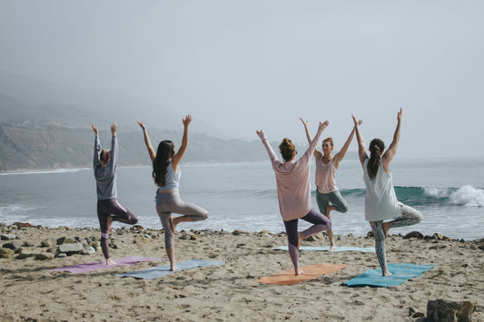 Fünf Frauen stehen am Meeresufer und machen Yoga